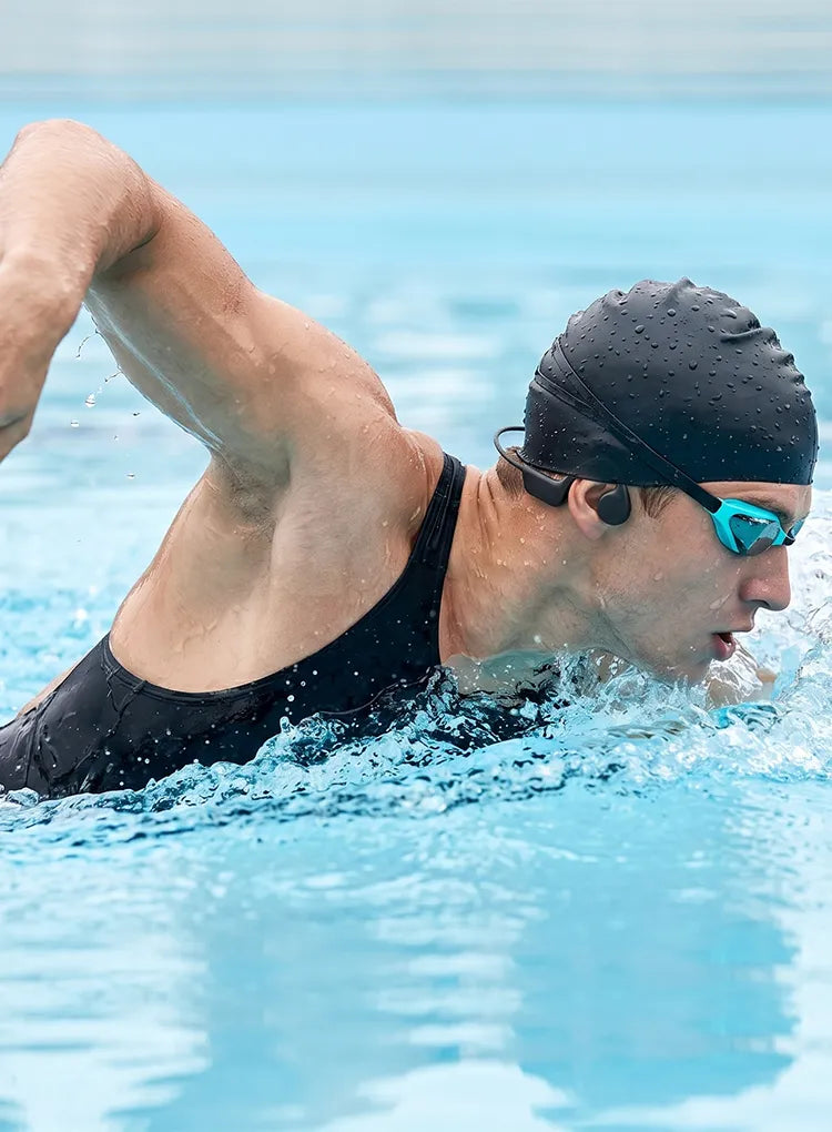 A swimmer in a black swimsuit, black swim cap, and blue-tinted goggles swimming in a bright blue pool.