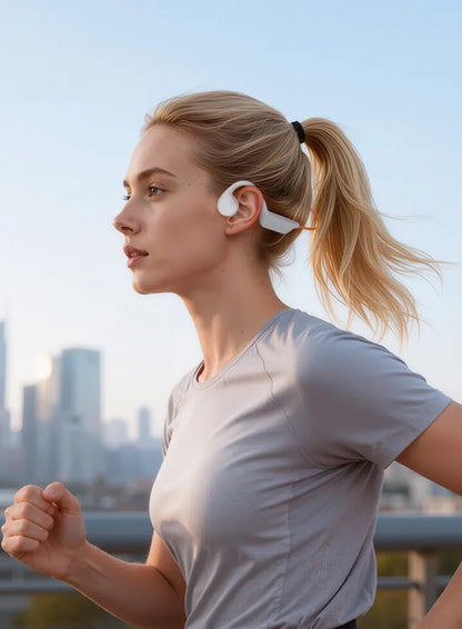 A woman jogging outdoors wearing a white over-ear wireless earbud behind her ear, with a blurred city skyline in the background.