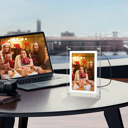 Laptop displaying a video call with people in festive attire, next to a transparent CaptiPix Digital Photo Frame on a table.