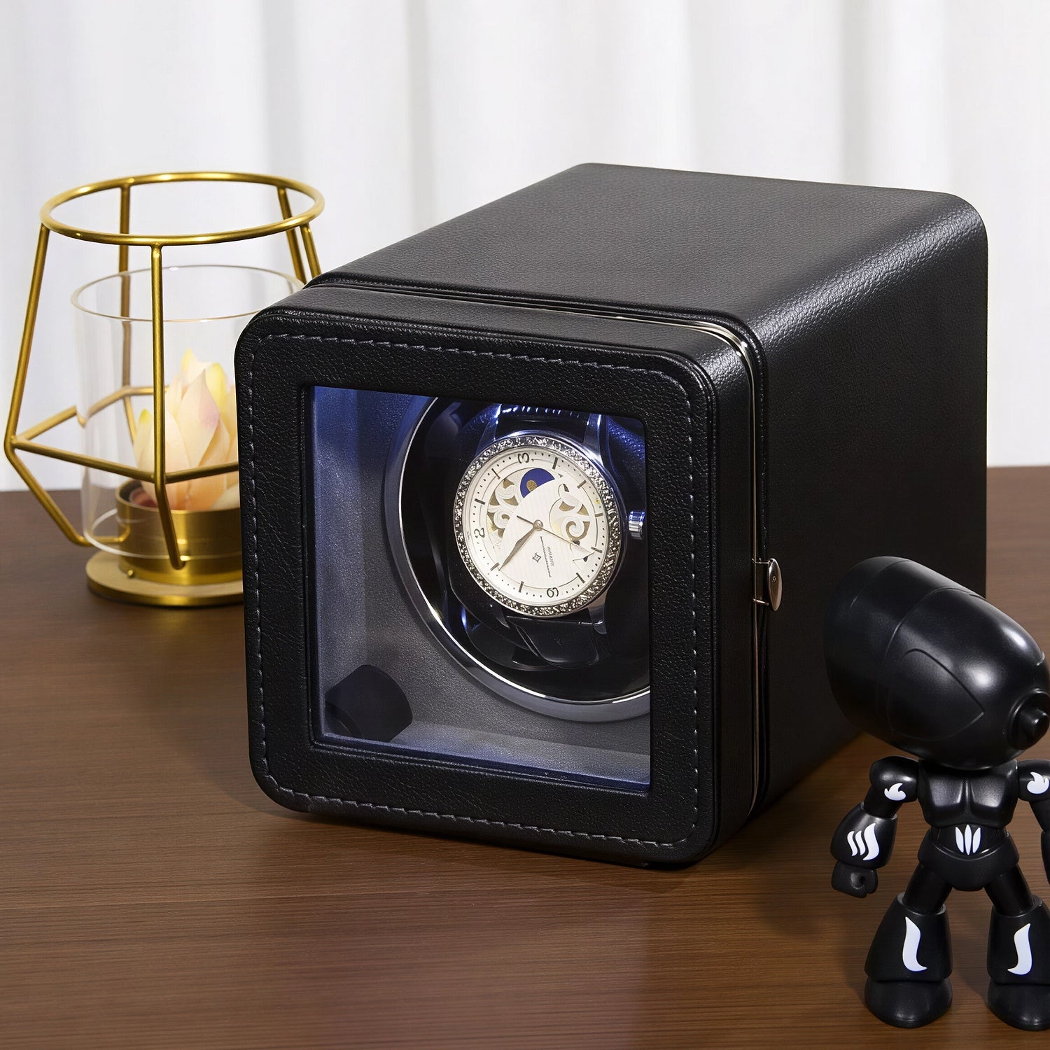 Black ChroniNova Watch Winder with a watch inside on a wooden surface, next to a small black figurine and gold candle holder.