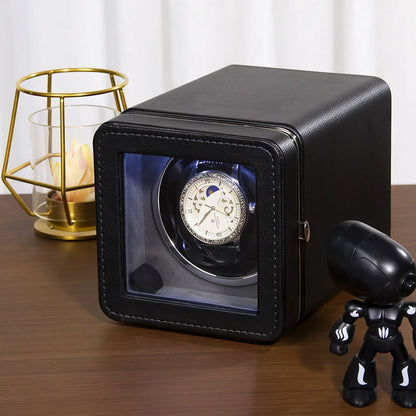 Black ChroniNova Watch Winder with a watch inside on a wooden surface, next to a small black figurine and gold candle holder.
