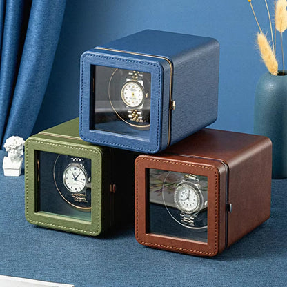 Three ChroniNova Watch Winder in blue, green, and brown on a blue surface with a decorative plant in the background.