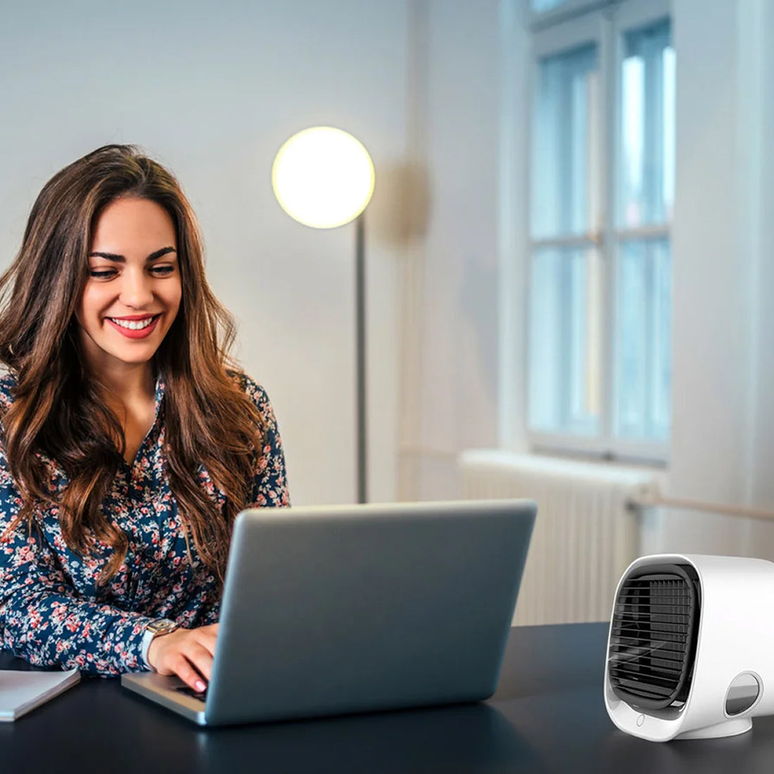 Woman using a laptop with a small Coolevax Portable Air Conditioner on a desk in a bright room.