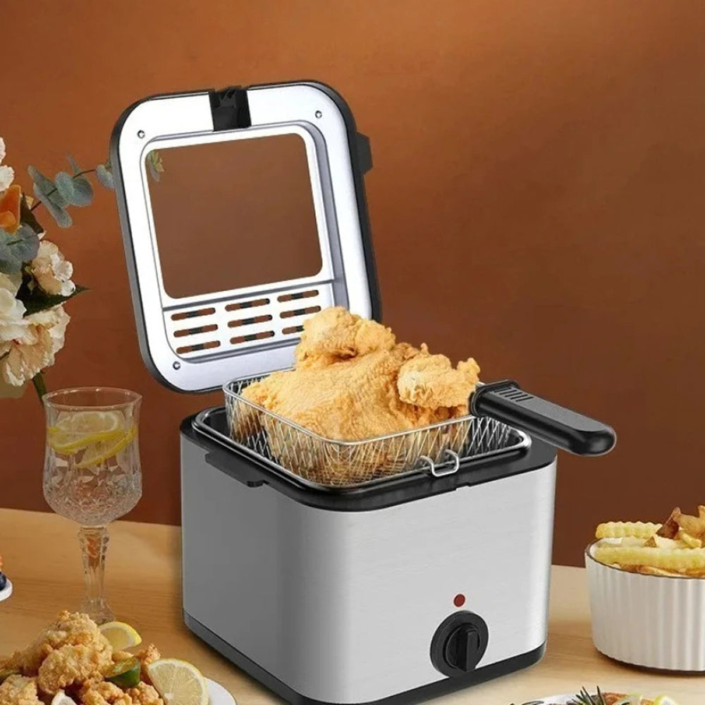 A countertop CryspiPro Fryer with its lid open, showing a golden-brown fried chicken basket filled with food. The fryer has a silver and black body, sits on a wooden table, and is surrounded by food items, a glass of lemonade, and flowers against a warm brown background.