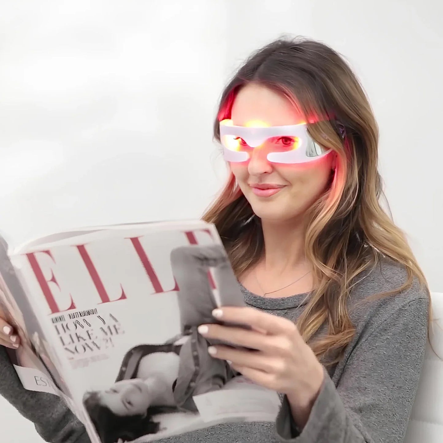 Woman wearing LED Eyviola Eye Massager and reading a magazine with a phone in her other hand.