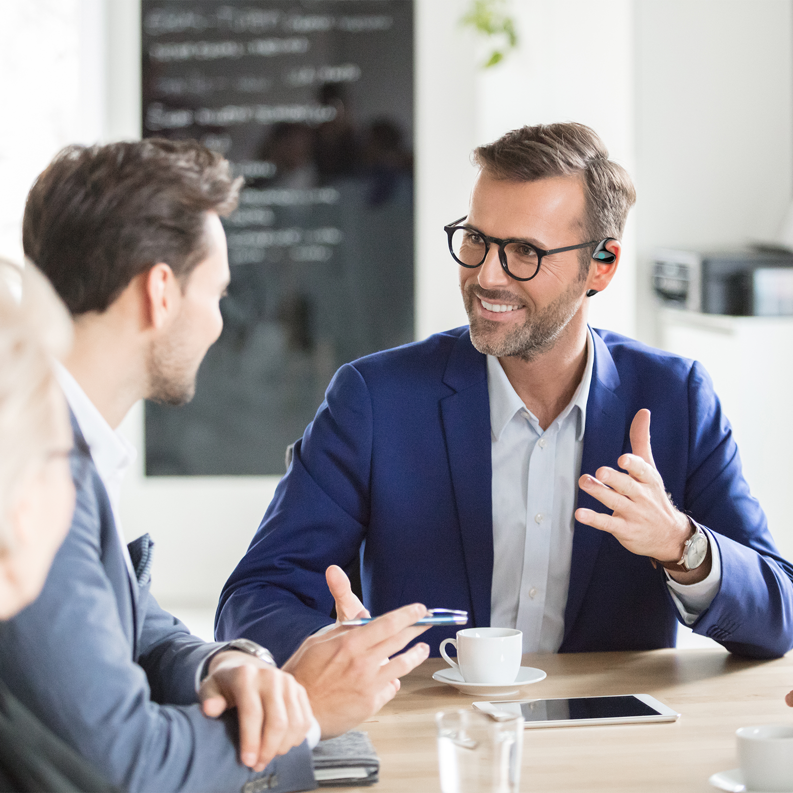 Two men in business attire sitting at a table with a tablet and coffee, engaged in a conversation.